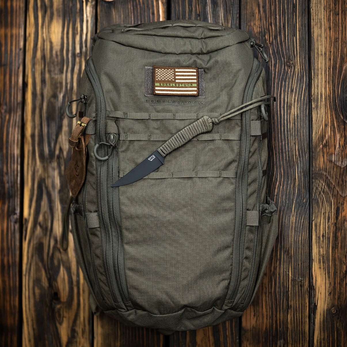 `Olivgrüner Eberlestock Tactical Rucksack mit MOLLE-System und Outdoor-Ausrüstung auf rustikalem Holzuntergrund`
